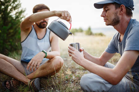 one man pours another tea into a mug from a kettle, sitting and relaxing after trekking. Traveler man in blue baseball cap holding a mug of tea after hiking. Travel peopleの写真素材