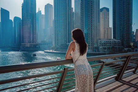 Happy beautiful tourist woman in fashionable summer white dress walking and enjoying in Dubai marina in United Arab Emirates. Luxury and comfortable tourism season in UAE. Warm summer day.の写真素材