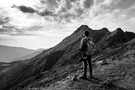 Young man standing on top of cliff in summer mountains at sunset and enjoying view of nature. The idea and concept of hiking, photography and travel. Russia, ski resort Rosa Khutor, Rosa Peakの写真素材
