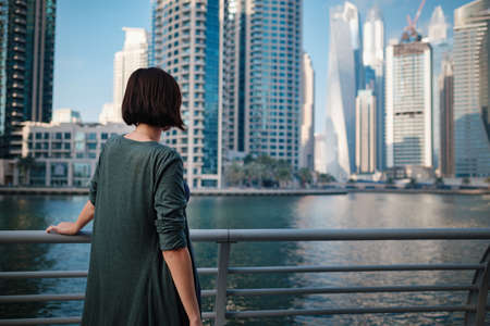 Happy young female traveler in the big city of Dubai, famous place Dubai marina. Luxury and comfortable tourism season in United Arab Emirates. Back or rear view of young woman in dressの写真素材
