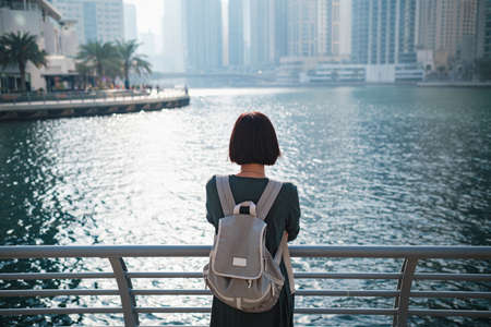 Happy young female traveler in the big city of Dubai, famous place Dubai marina. Luxury and comfortable tourism season in United Arab Emirates. Back or rear view of young woman in dress and backpackの写真素材