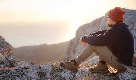 A young man standing on top cliff in spring mountains at sunset and enjoying view of nature. Mountain and coastal travel, freedom and active lifestyleの写真素材
