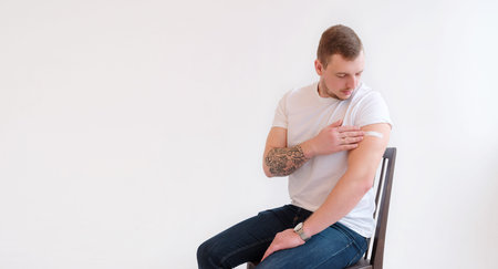 Man in white T-shirt smiling after receiving vaccination on white background. Man showing his arm...