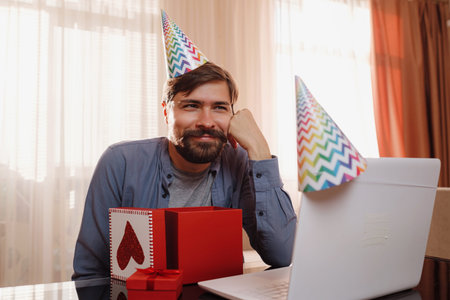 happy young man sitting at the table and talking by video call to friends. A man opens his gift,...