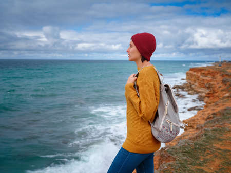 Hipster woman traveler with backpack in yellow sweater and red hat enjoying wind on the coast. storm and thunderstorms in the distance over the sea, the happiness and freedom of travel and discoveryの写真素材