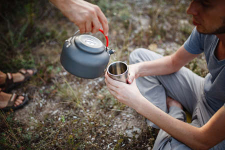 one man pours another tea into a mug from a kettle, sitting and relaxing after trekking. Traveler man in blue baseball cap holding a mug of tea after hiking. Travel peopleの写真素材