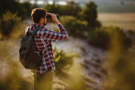 Guy looking at binoculars in hill. man in t-shirt with backpack. Young Caucasian man during hike in valley landscapeの写真素材