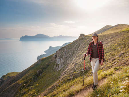 Female hiker walking on dirt road. Woman using trekking poles during hike. enjoying a sunny view of the sea. Mountain and coastal travel, freedom and an active lifestyleの写真素材