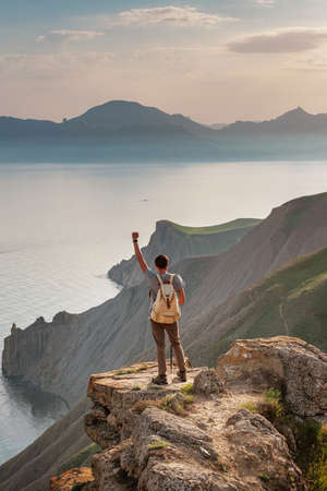 Young man travels alone on the backdrop of the mountains standing on top of a mountain with raised hands, the lifestyle concept of traveling outdoors.の写真素材