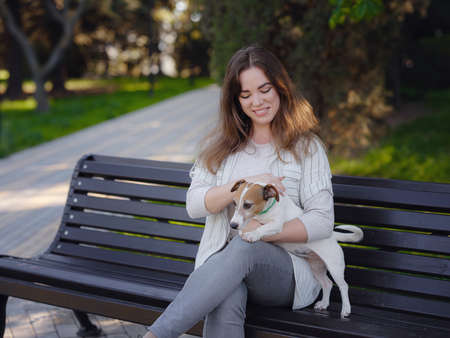 Young woman with her cute Jack Russell Terrier outdoor. Lovely pet. A nice spring evening walk in a green park hugging on a benchの写真素材