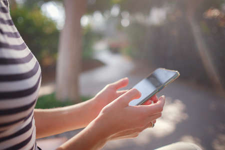 A woman wearing a protective mask looks at her smartphone display at park. Tourist female hands holding a gadget against a blurry summer backgroundの写真素材