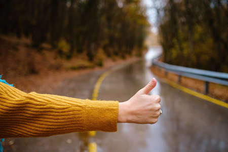 Close up shot of a womans hand doing hitchhiking sign near the country roadside during vacation trip in mountains fall forestの写真素材
