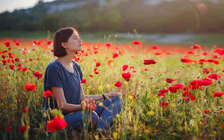 A beautiful woman meditates on a poppy field at sunset. Wellness well-being happiness concept.の写真素材