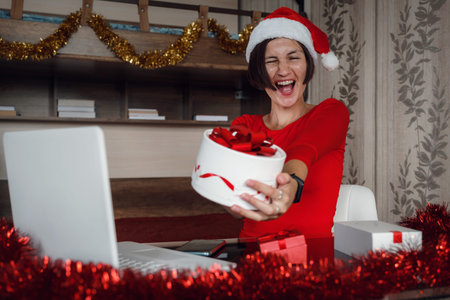 A happy woman in a red shirt is sitting in front of a computer screen with a gift box in her hands....