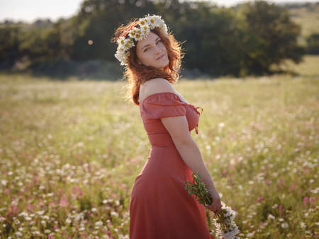 red-haired woman wearing dress and chamomile wreath on field on summer evening. concept of digital detox, digital cleanse, reconnecting with nature, calmness and silenceの写真素材