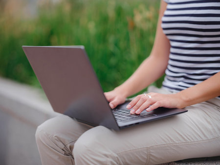 happy asian female student working with laptop in modern university campus park. woman in striped T-shirt and light trousers works at gray laptop. Hands closeupの写真素材