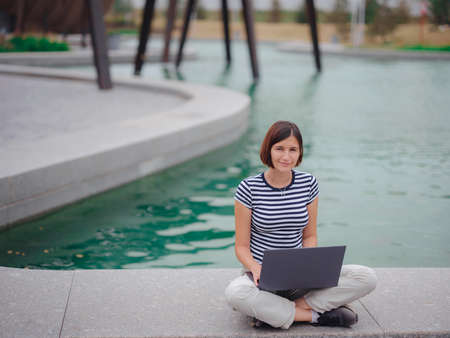 happy asian female student working with laptop in modern university campus park. woman in striped T-shirt and light trousers typing at gray laptop against the backdrop of a pond.の写真素材