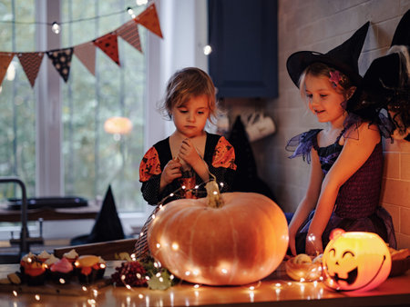 little sisters in witch costume playing with glowing jack-o-lantern during Halloween celebration in dark kitchen room, excited kids and pumpkins on tableの写真素材