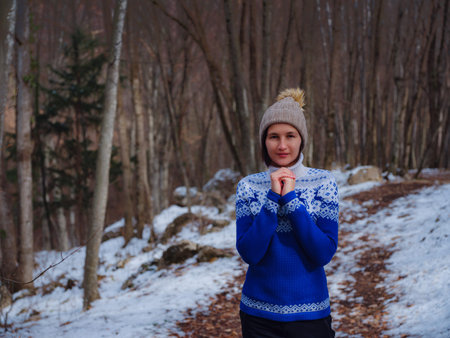 Beautiful woman standing among trees in winter forest. Wearing hat and blue sweater. Caucasian Asian...