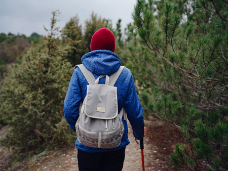 Portrait of woman hiker in blue coat and red hat with hiking stick standing among juniper bushes, cloudy winter sky on background. Travel and active lifestyle concept.の写真素材