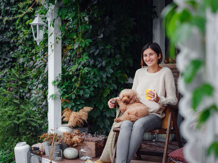 Young asian woman resting outdoors with cup of coffee on porch of country house, chilling outside with poodle dog. autumn lifestyle, leisure free time concept. Copy spaceの写真素材