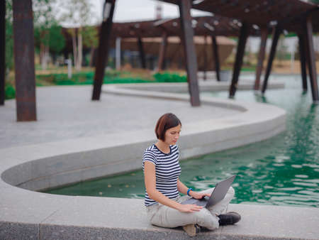 happy asian female student working with laptop in modern university campus park. woman in striped T-shirt and light trousers typing at gray laptop against the backdrop of a pond.の写真素材