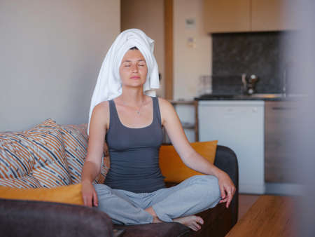 woman sitting on sofa, lotus position puts hands on knees, closed eyes, enjoy meditation, do yoga. No stress, healthy habits, conscious lifestyle, anxiety relief conceptの写真素材