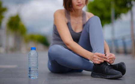 outdoor sports, workout and wellness concept. fitness woman tying shoelaces sneakers on road. cheerful runner sits on floor on streets of city. Active Hispanic woman tying lace shoes before running.の写真素材