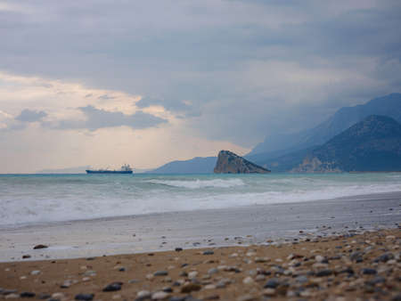 Cargo ship in calm blue Mediterranean in Anatalya. Traveling along the Mediterranean Sea, view of the rocky coast of Antalya. In distance, cargo ships are visible.の写真素材