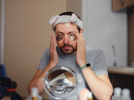 patches for eyes. Young man in kitchen with headband on his head and mirror apply use under eye...