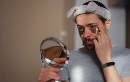 patches for eyes. Young man in kitchen with headband on his head and mirror apply use under eye anti-wrinkle patches, men do morning facial procedures with hydrogel beauty product, skincare.の写真素材