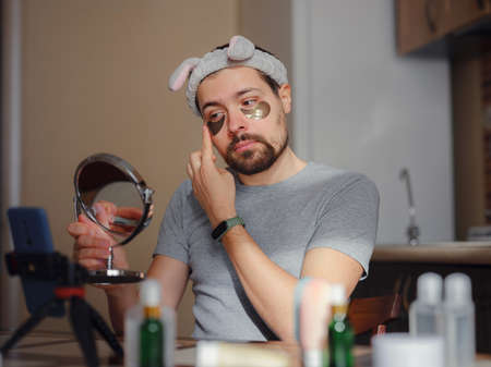 patches for eyes. Young man in kitchen with headband on his head and mirror apply use under eye anti-wrinkle patches, men do morning facial procedures with hydrogel beauty product, skincare.の写真素材