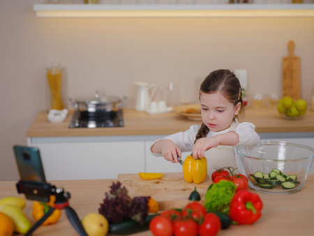 Young Girl Vlogger Making Social Media Video About Cooking At Home, looking on smartphone on tripod. episode about healthy eating. concept of attraction children to a healthy diet of early ageの写真素材