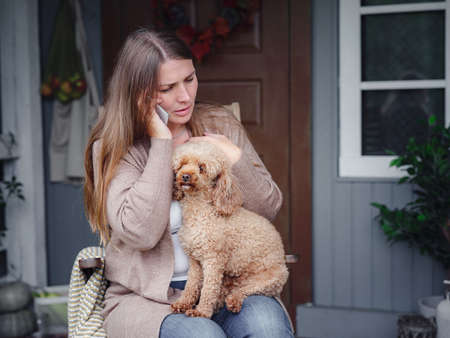 middle aged caucasian woman in brown cardigan sits on terrace of house with her beige poodle and emotionally speaks with friend via smartphone. morning good and bad news, autumn lifestyle conceptの写真素材