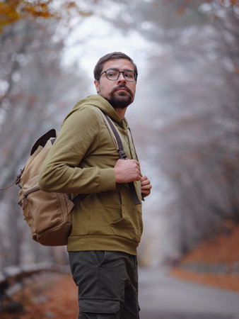 Young handsome man posing in autumn forest on road. young hipster guy with backpack , traveler...