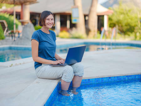Young business woman working at computer by hotel pool. Young lady downshifter working at laptop and enjoys and relaxed environment, working day. Online freelance work on vacation.の写真素材