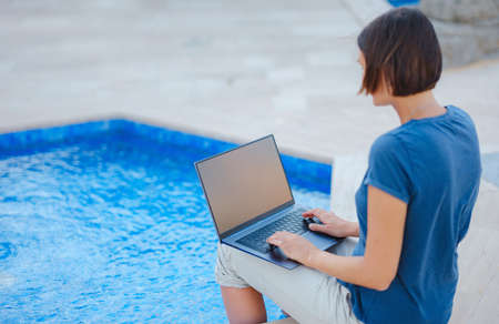 Young business woman working at computer by hotel pool. Young lady downshifter working at laptop and enjoys and relaxed environment, working day. Online freelance work on vacation.の写真素材
