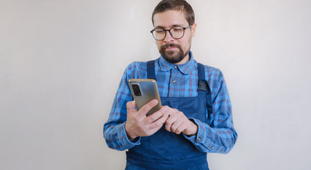 young man in blue work suit doing repair apartment. home renovation concept. Guy with smartphone texting and tapping on cellphone, searching for solution on Internet, ordering goods for repairsの写真素材