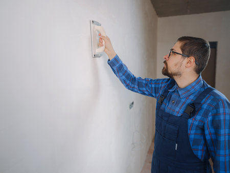 young man in blue work suit doing repair apartment. home renovation concept. Use sandpaper to sand wall surface. Surface leveling, work inside house.の写真素材