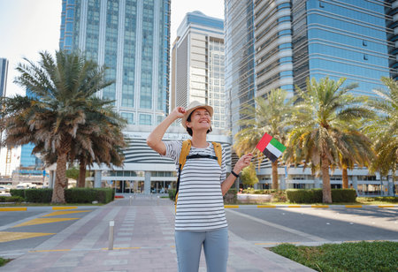 Happy young asian female traveler with backpack and hat with UAE flag against scenic skyscrapers in Abu Dhabi. Immigration, student education and arab emirates citizenship conceptの写真素材
