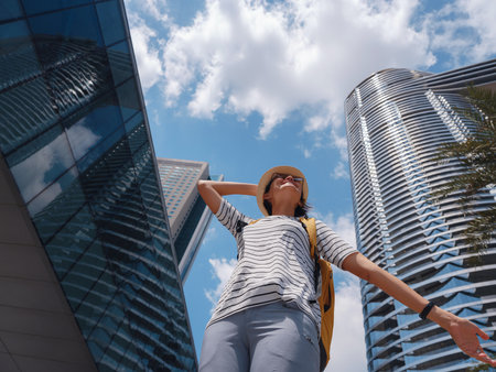 travel to the United Arab Emirates, Happy young asian female traveler with backpack and hat in big city of Abu Dhabi, skyscrapers bottom view. Vacation and tourist destination concept.の写真素材