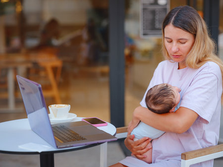 Diverse people portrait. Creative businesswoman balancing work and motherhood. Beautiful young mother working with laptop computer and breastfeeding, holding and nursing her newborn baby at cafe.の写真素材