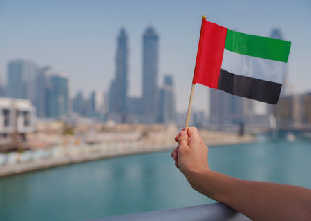 Close up of a female hand holding tiny flag of UAE against Dubai skyline. Photo of UAE national day celebration spirit.の写真素材