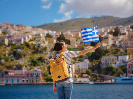 Nice asian Happy Female with backpack anf greek flag Enjoying her holidays on Symi Islands. View of port Symi or Simi, is tiny island of Dodecanese, Greece, calm atmosphere and fabulous architecture.の写真素材