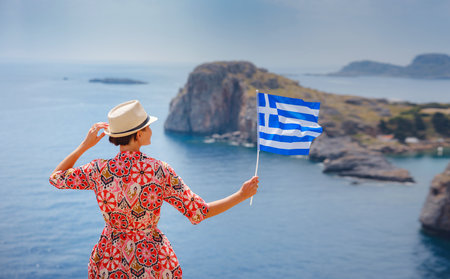 Nice Happy Female Enjoying Sunny Day on Greek Islands. Travel to Greece, Mediterranean islands outside tourist season. Young traveling woman with national greek flag enjoying view on seaの写真素材