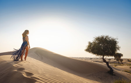 Desert adventure. Young Arabian Woman in sexy boho blue makrame dress in sands dunes of UAE desert at sunset. The Dubai Desert Conservation Reserve, United Arab Emirates.の写真素材