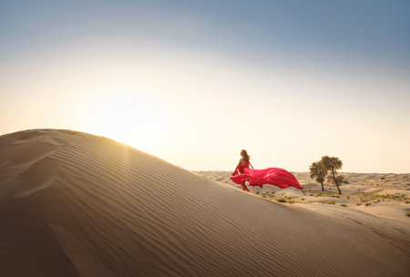 Desert adventure. Young arabian Woman in red silk dress in sands dunes of UAE desert at sunset, fantastic view. The Dubai Desert Conservation Reserve, United Arab Emirates.の写真素材