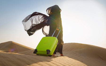 Arabian woman in traditional abaya costume with bright light green or yellow suitcase in sands. Concept and idea of travel to United Arab Emirates, sand dunes in desert of Dubai at sunset.の写真素材