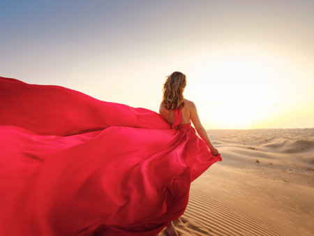 Desert adventure. Young arabian Woman in red silk dress in sands dunes of UAE desert at sunset, fantastic view. The Dubai Desert Conservation Reserve, United Arab Emirates.の写真素材