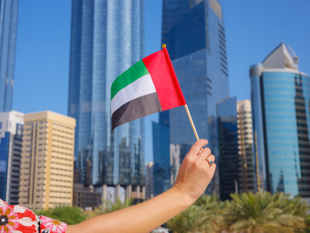 Close up of female hand holding tiny flag of UAE against Abu Dhabi skyline. Photo of UAE national day celebration spirit.の写真素材